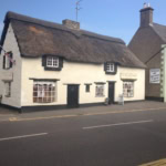 A traditional, white-painted thatched cottage with a thick, shaggy straw roof stands beside a street. A sign on the building says "The Thatched Cottage" and a small Union Jack flag hangs from an upstairs window.
