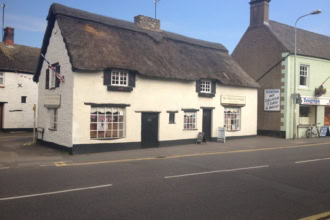 A traditional, white-painted thatched cottage with a thick, shaggy straw roof stands beside a street. A sign on the building says "The Thatched Cottage" and a small Union Jack flag hangs from an upstairs window.