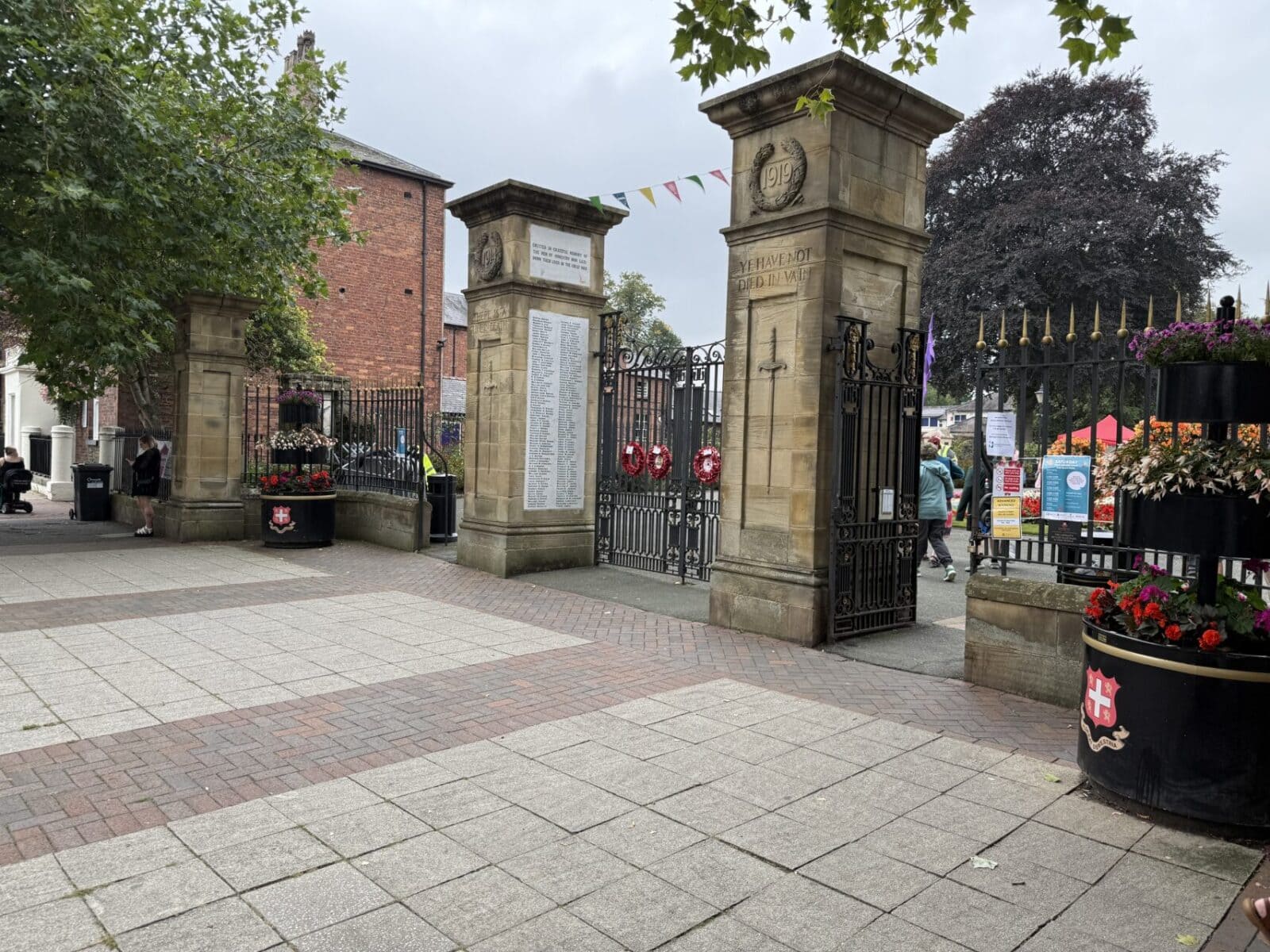 Stone entrance pillars with war memorial plaques and wrought iron gates leading into Cae Glas Park, with colorful bunting overhead, flower planters, and brick buildings in background