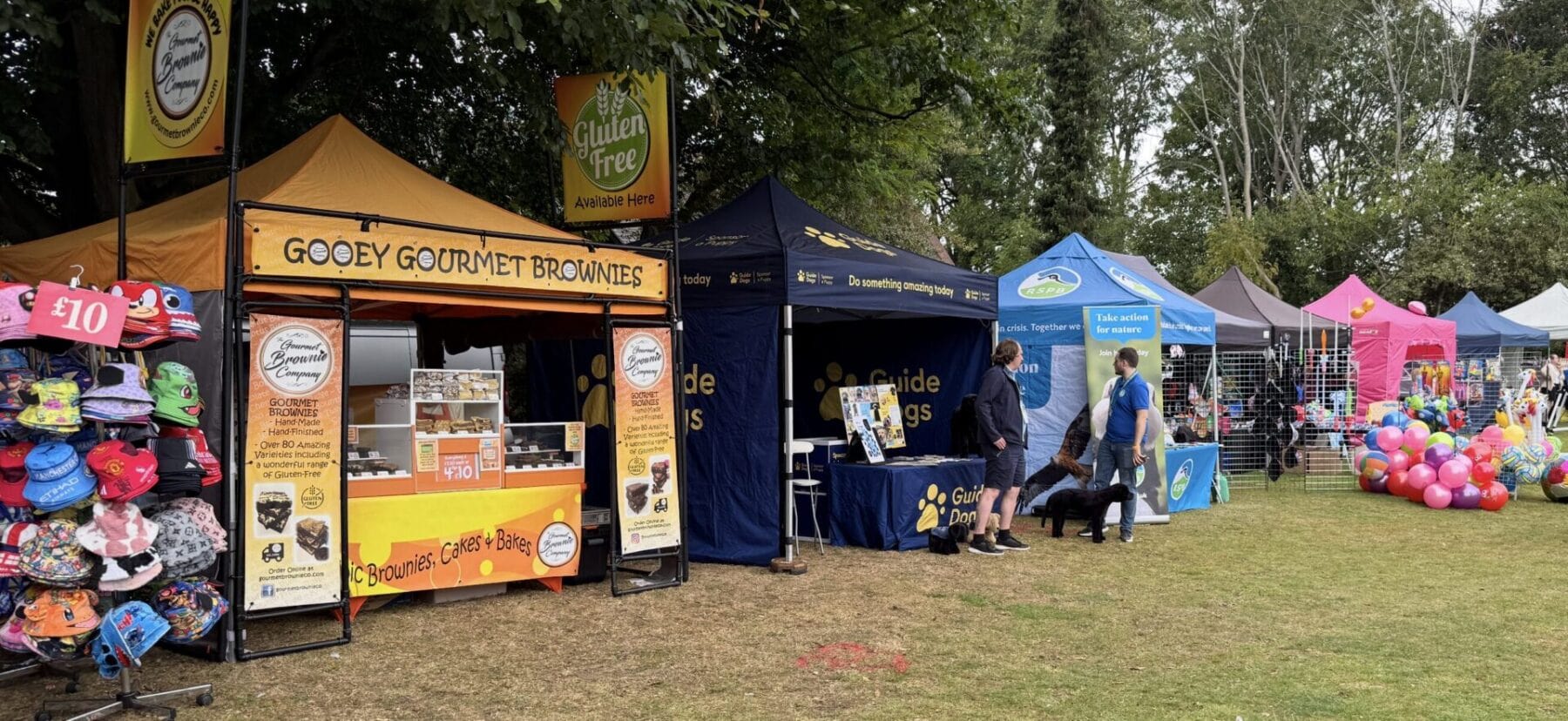 Alt Text: Market stalls and vendor tents at Oswestry Balloon Festival with colorful merchandise displays, food vendors, and visitors browsing on grass field surrounded by trees