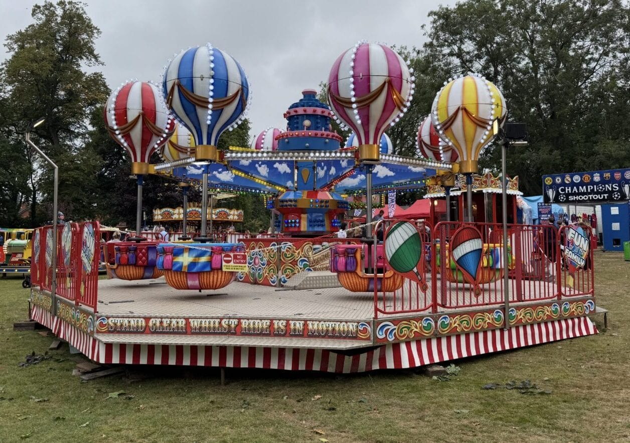 Colorful carnival ride with hot air balloon-themed gondolas in red, blue, pink and yellow stripes at children's funfair with safety barriers and other rides in background