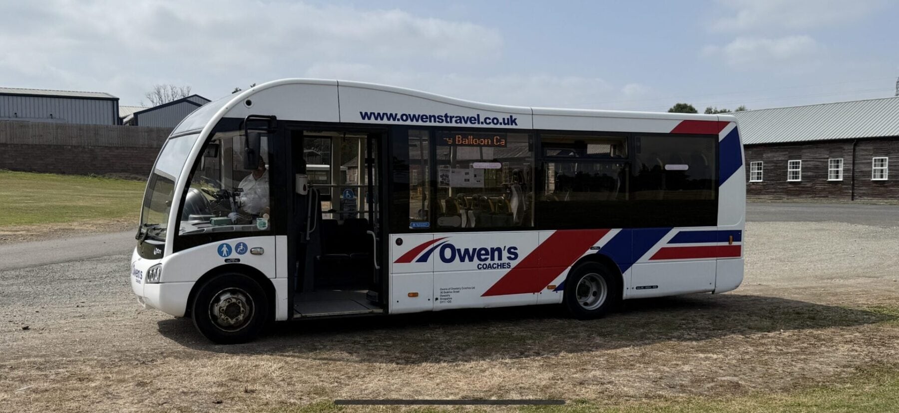 White Owen's Coaches bus with red and blue diagonal stripes parked on gravel, displaying "Balloon L3" destination and website www.owenstravel.co.uk, with industrial buildings in background