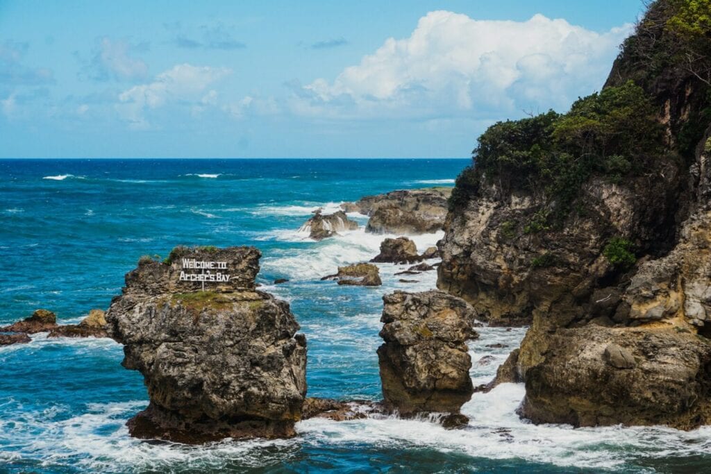 Rocky coastline at Archer's Bay with dramatic limestone cliffs, turquoise ocean water, white foam waves crashing against jagged rocks, and "Welcome to Archer's Bay" sign on cliff face