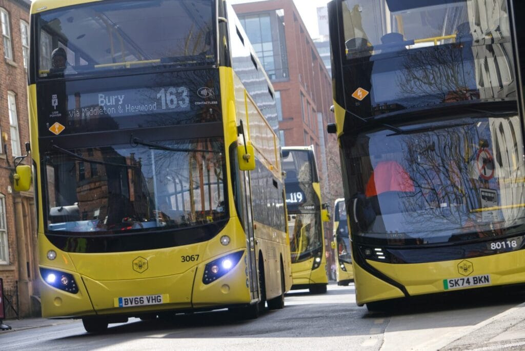 Two yellow Bee Network buses (numbers 3067 and 80118) on a Manchester street, with route 163 visible on the front bus display