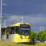 Yellow and grey Metrolink tram number 3083 displaying "Eccles" destination, stopped at a platform with overhead power lines and passengers nearby