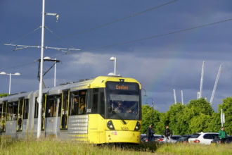 Yellow and grey Metrolink tram number 3083 displaying "Eccles" destination, stopped at a platform with overhead power lines and passengers nearby