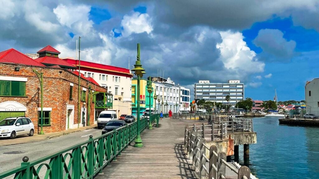 Scenic waterfront in Bridgetown, Barbados with a boardwalk, colorful colonial buildings, parked cars, and people strolling under a partly cloudy sky.