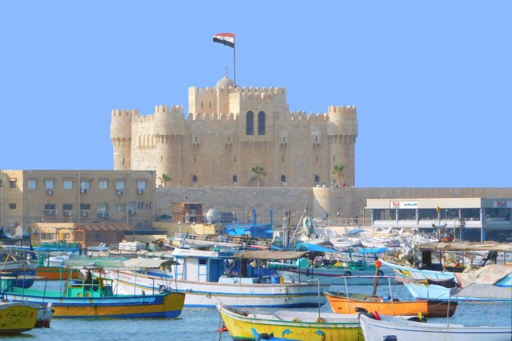 Colorful fishing boats in Alexandria harbor with historic Citadel of Qaitbay fortress in background, Egyptian flag flying from castle tower against blue sky