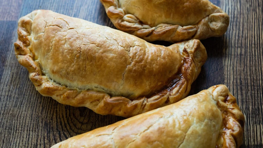 Three freshly baked Cornish pasties on a wooden surface, representing British cuisine featured in the United Kingdom travel guide.