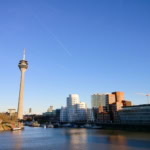 The skyline of Düsseldorf, Germany, features the Rheinturm television tower and several modern buildings along the banks of the Rhine River. The water is calm and reflects the buildings, and the sky is a clear blue with contrails.