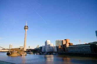 The skyline of Dรผsseldorf, Germany, features the Rheinturm television tower and several modern buildings along the banks of the Rhine River. The water is calm and reflects the buildings, and the sky is a clear blue with contrails.