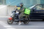 Uber Eats courier on a scooter waiting at a city crossing, wearing a helmet and carrying a bright green delivery bag.