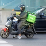Uber Eats courier on a scooter waiting at a city crossing, wearing a helmet and carrying a bright green delivery bag.