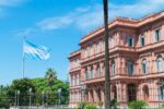 Pink neoclassical Casa Rosada presidential palace in Buenos Aires with Argentine flag flying and palm trees under blue sky