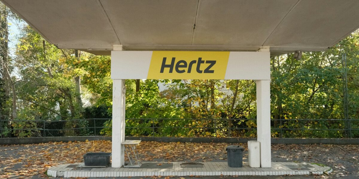 Hertz car rental pickup canopy with distinctive yellow and black branding in tree-lined location with autumn foliage