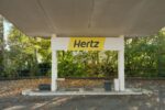 Hertz car rental pickup canopy with distinctive yellow and black branding in tree-lined location with autumn foliage