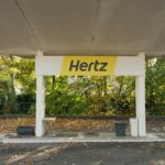Hertz car rental pickup canopy with distinctive yellow and black branding in tree-lined location with autumn foliage