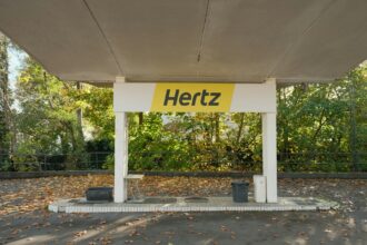 Hertz car rental pickup canopy with distinctive yellow and black branding in tree-lined location with autumn foliage