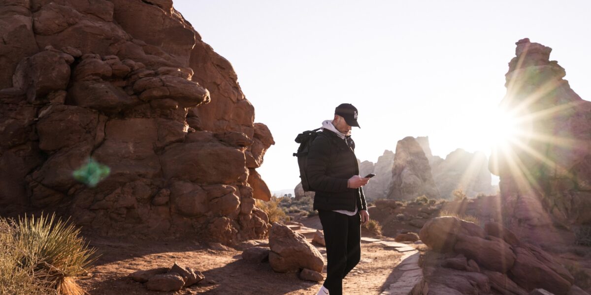 Hiker with backpack checking mobile device among red rock formations in desert landscape with sun rays