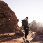Hiker with backpack checking mobile device among red rock formations in desert landscape with sun rays
