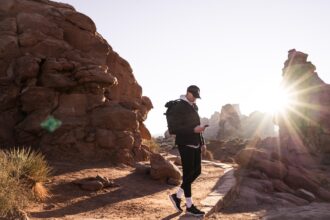 Hiker with backpack checking mobile device among red rock formations in desert landscape with sun rays