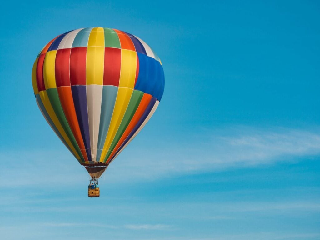 A colorful hot air balloon, with a pattern of vertical stripes in various colors like red, yellow, green, and blue, floats high against a clear, vibrant blue sky.