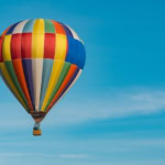 A colorful hot air balloon, with a pattern of vertical stripes in various colors like red, yellow, green, and blue, floats high against a clear, vibrant blue sky.