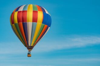 A colorful hot air balloon, with a pattern of vertical stripes in various colors like red, yellow, green, and blue, floats high against a clear, vibrant blue sky.