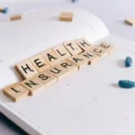 Wooden scrabble tiles spelling ‘Health Insurance’ on a calendar with blue pills, symbolising travel insurance preparation and medical coverage planning
