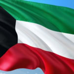 Kuwait flag with green, white, red horizontal stripes and black triangle waving against blue sky