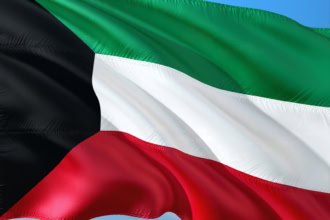 Kuwait flag with green, white, red horizontal stripes and black triangle waving against blue sky