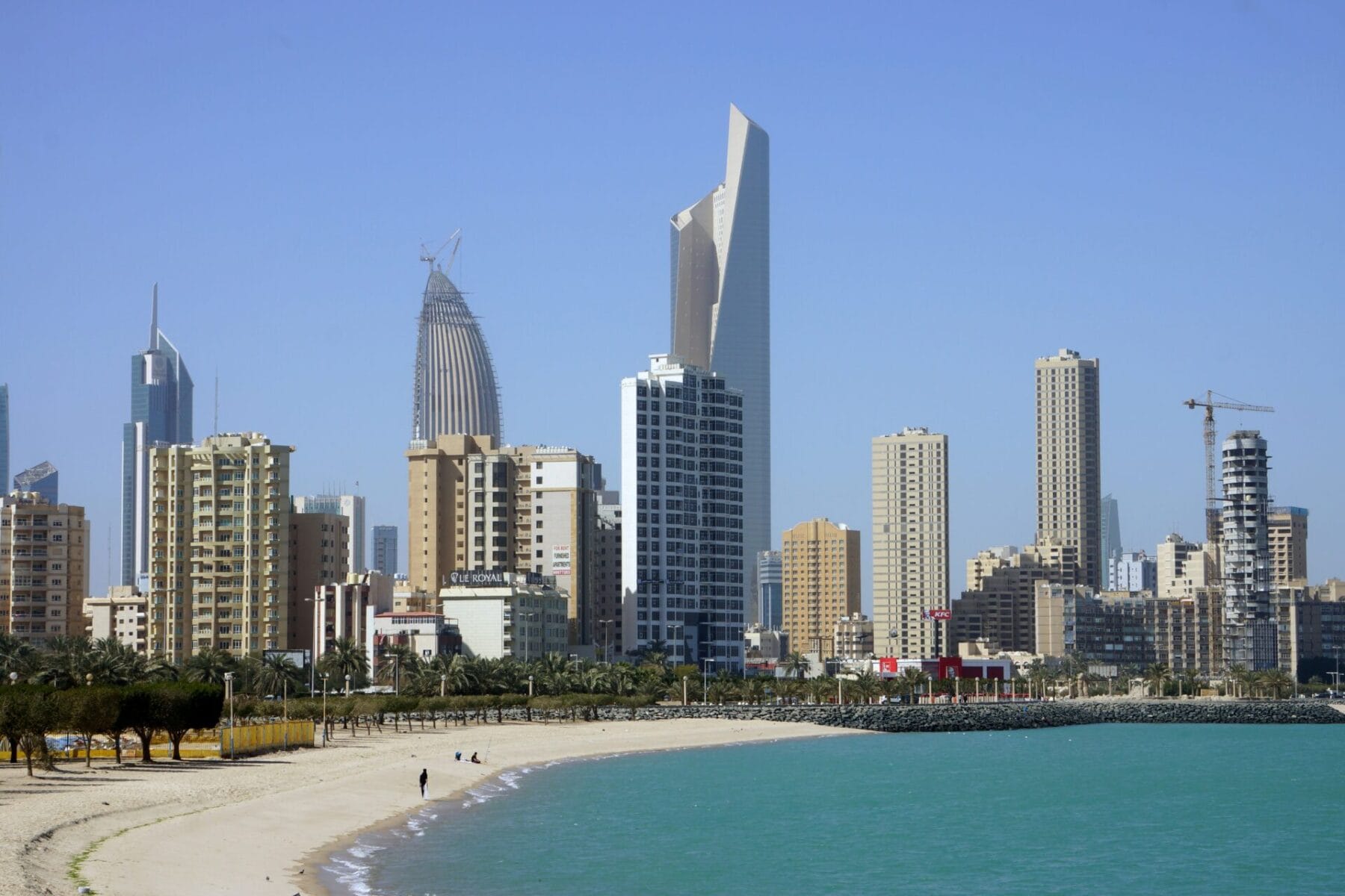 Modern Kuwait City skyline featuring contemporary skyscrapers and high-rise buildings viewed from a pristine beach with turquoise waters and white sand, palm trees in foreground.