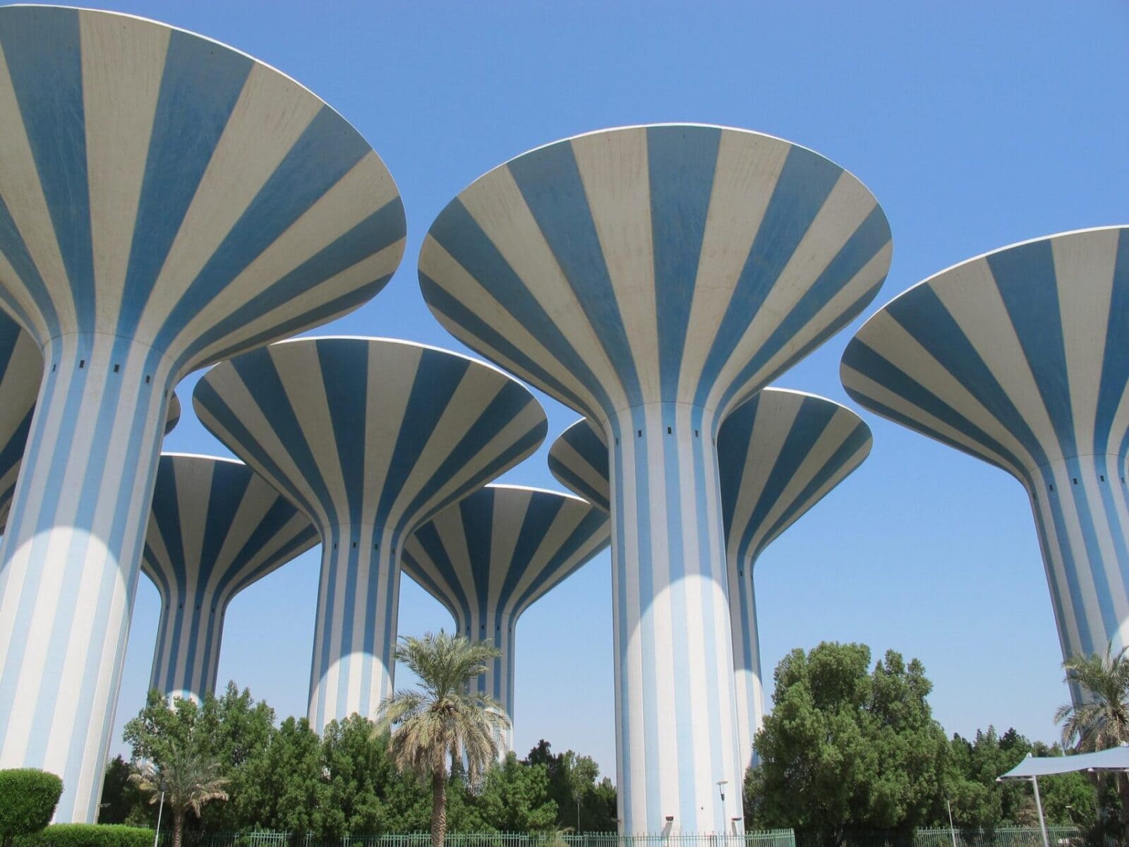 Three distinctive mushroom-shaped water towers with blue and white striped design against clear blue sky, surrounded by palm trees and landscaped gardens in Kuwait.