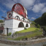The Great Laxey Wheel, a large red and white painted waterwheel housed in a stone building with arched openings, set in a green valley surrounded by trees and stone ruins under a dramatic cloudy blue sky.