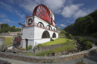 The Great Laxey Wheel, a large red and white painted waterwheel housed in a stone building with arched openings, set in a green valley surrounded by trees and stone ruins under a dramatic cloudy blue sky.