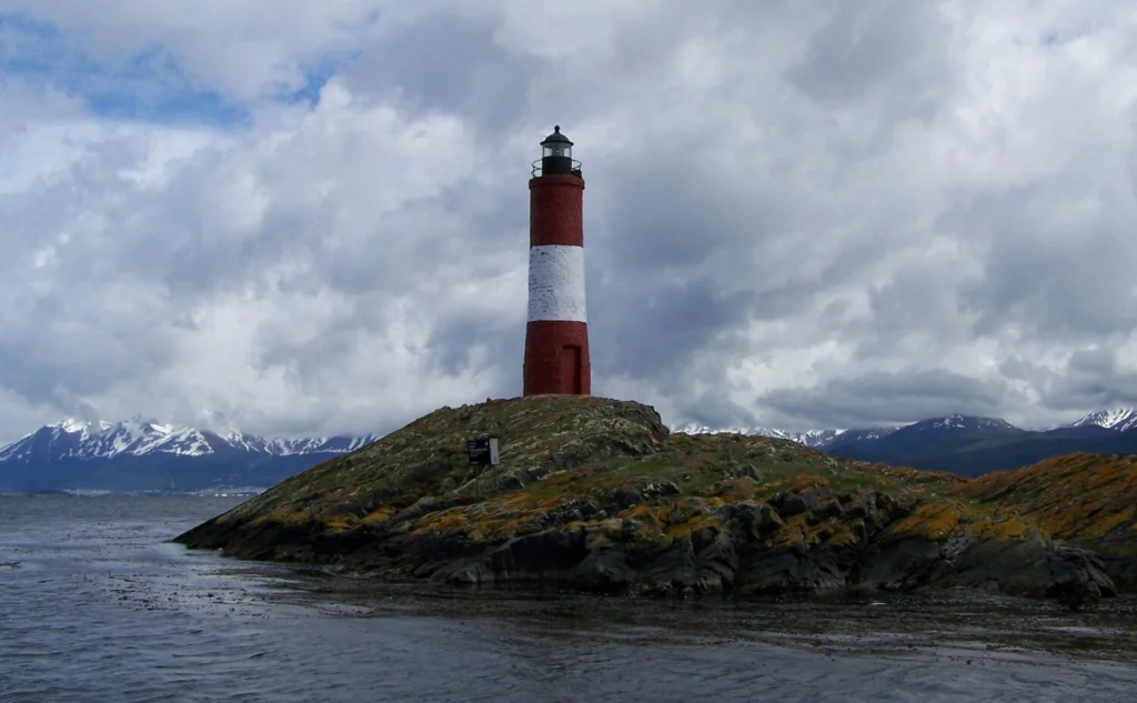 Red and white lighthouse on a rocky island near Ushuaia, with snow-capped mountains in the background.
