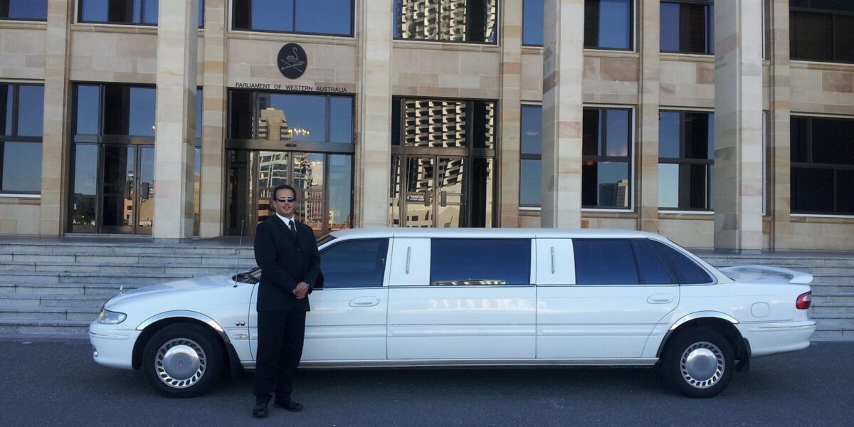 Chauffeur in black suit standing beside white stretch limousine outside Parliament of Western Australia building