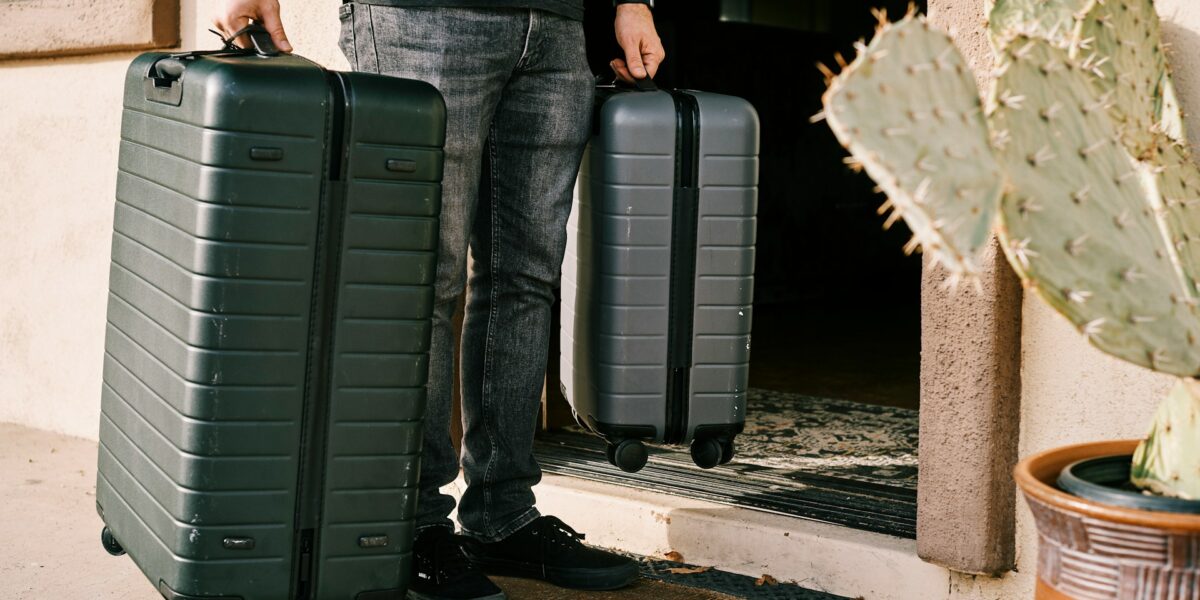 Person with tattooed arm pulling two dark green hard-shell suitcases outside building entrance with cactus plants