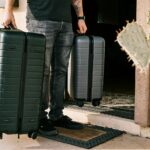 Person with tattooed arm pulling two dark green hard-shell suitcases outside building entrance with cactus plants