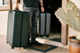 Person with tattooed arm pulling two dark green hard-shell suitcases outside building entrance with cactus plants