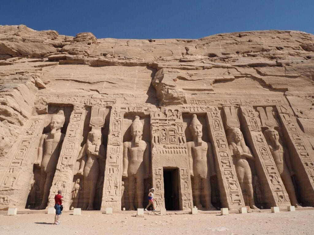 Facade of Abu Simbel temple with four massive seated statues of Pharaoh Ramesses II carved into sandstone cliff, tourists for scale, hieroglyphs covering temple walls