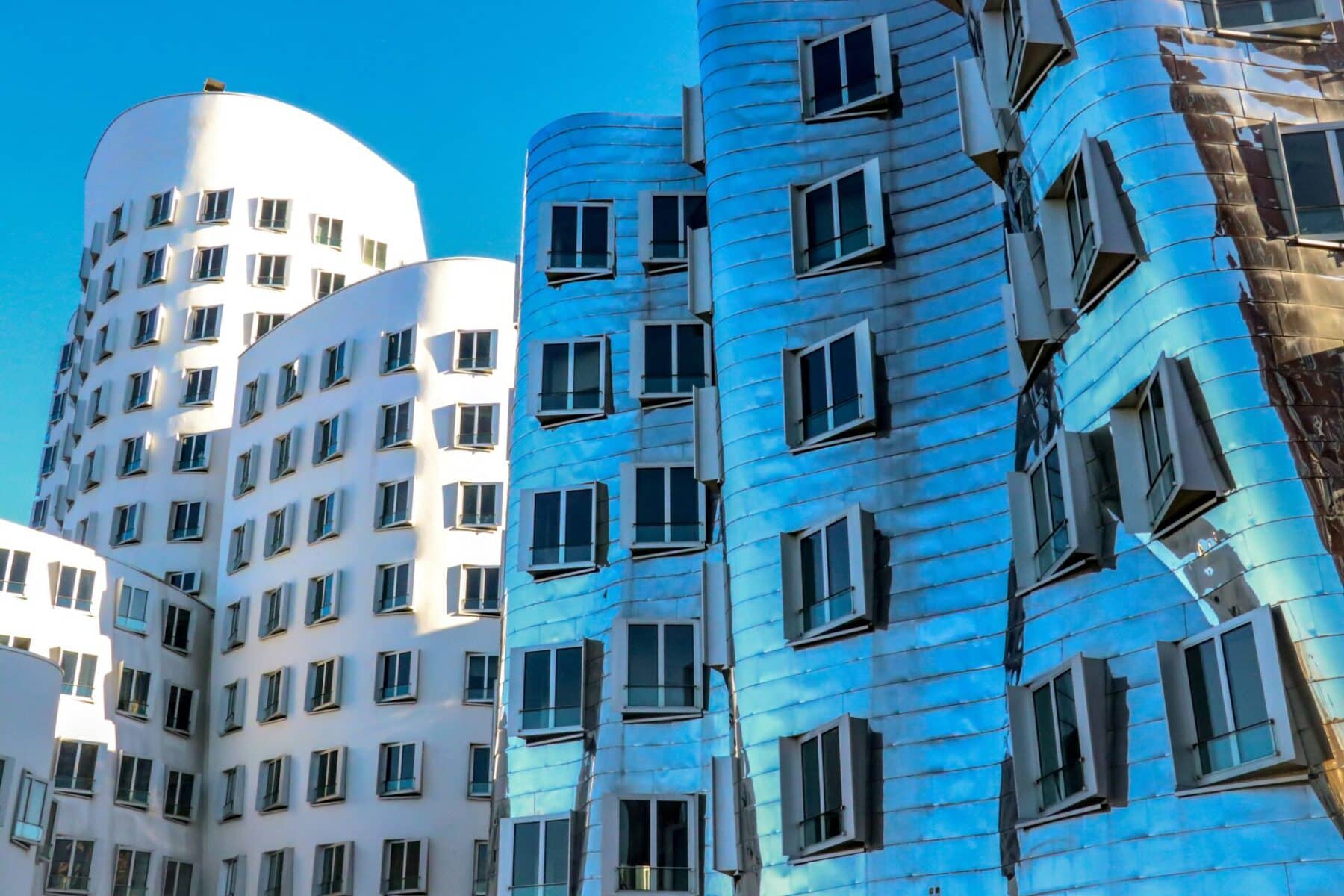 Close-up shot of several modern, curved buildings in the Media Harbour (MedienHafen) of Düsseldorf. The building on the right is covered in a shiny, metallic, and wrinkled facade, reflecting a blue sky, while the buildings behind it are white with a more standard design.