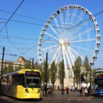 Yellow Manchester Metrolink tram and blue double-decker bus in city center with large white observation wheel and overhead electric cables against blue sky