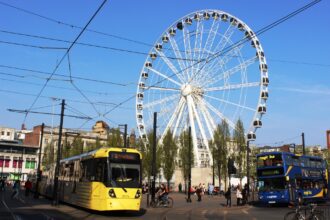 Yellow Manchester Metrolink tram and blue double-decker bus in city center with large white observation wheel and overhead electric cables against blue sky
