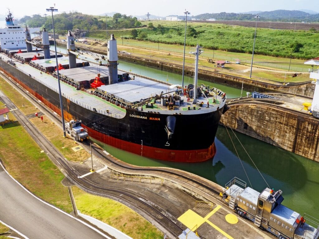 Cargo ship "LOUISIANA MAMA" passing through a lock in the Panama Canal, guided by machinery and surrounded by lush greenery.