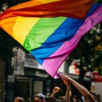 A person's arms are raised, holding up a large, flowing rainbow pride flag above a crowd. The flag's vibrant colors fill a significant portion of the frame, with blurred city buildings and trees in the background.