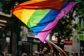 A person's arms are raised, holding up a large, flowing rainbow pride flag above a crowd. The flag's vibrant colors fill a significant portion of the frame, with blurred city buildings and trees in the background.