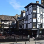 Two historic timber-framed buildings, a pub and a restaurant, stand side-by-side in a paved square. The building on the right, "The Shambles," is black and white, while the one on the left, "The Old Wellington," is a dark wood and white design. Outdoor seating is visible in front of both.