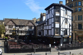 Two historic timber-framed buildings, a pub and a restaurant, stand side-by-side in a paved square. The building on the right, "The Shambles," is black and white, while the one on the left, "The Old Wellington," is a dark wood and white design. Outdoor seating is visible in front of both.