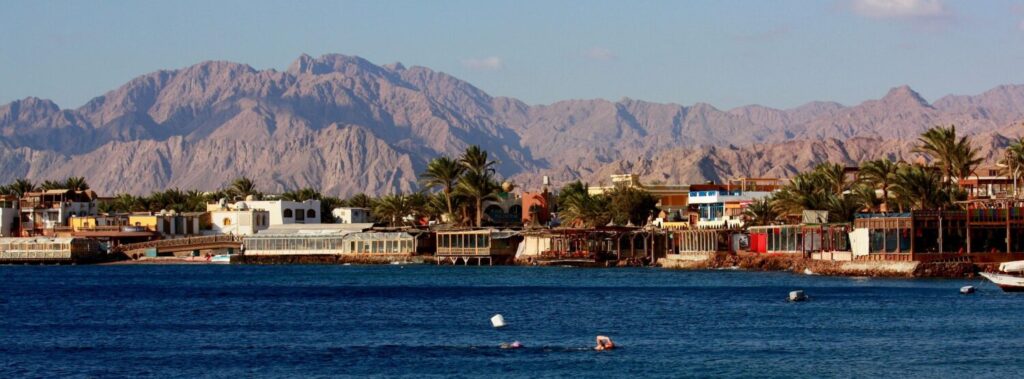 Coastal town of Sharm El-Sheikh with swimmers in the sea, palm trees, and mountains in the background.